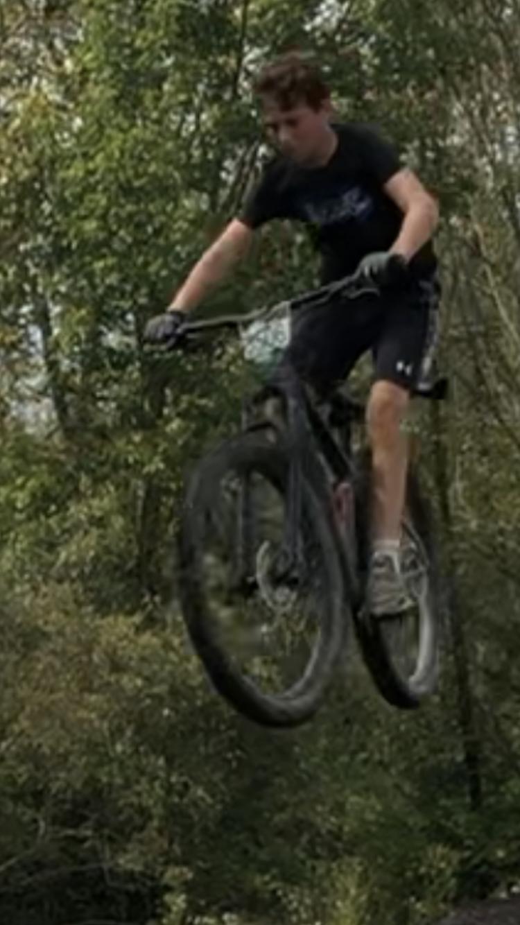 Trek Roscoe 8: A young person performing a jump on a mountain bike in a wooded area, with their feet off the pedals and both wheels in the air over a dirt ramp.