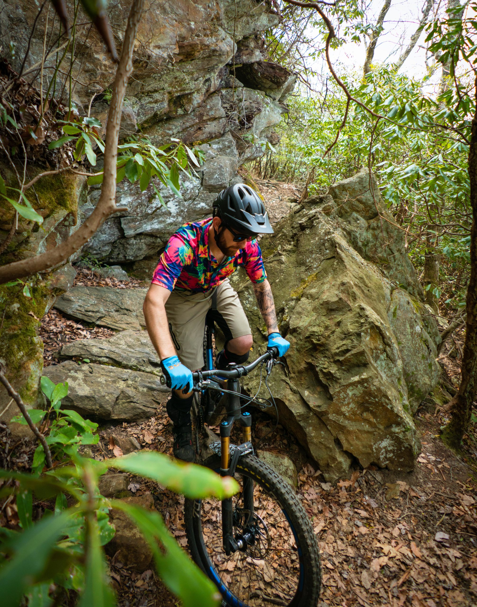 A mountain biker navigating a rocky trail surrounded by dense greenery. The rider is wearing a colorful floral shirt and gloves, along with a helmet, and is focusing on maneuvering through the rugged terrain. Laurel Mountain mountain bike trail.