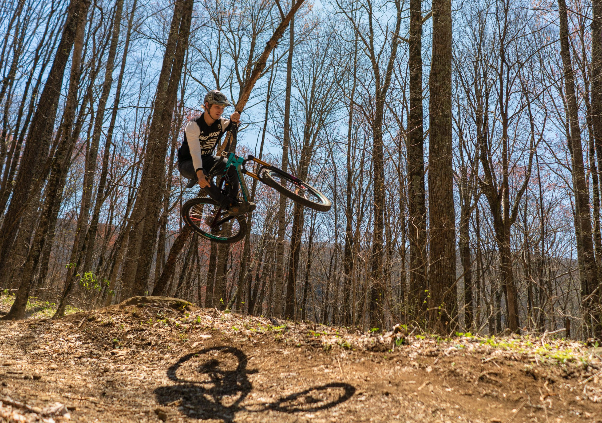 A person wearing a helmet and a long-sleeve shirt performs a jump on a mountain bike in a forested area. Tall trees and a clear blue sky are visible in the background, while the ground shows dirt and patches of greenery. The biker is mid-air, showcasing a dynamic action pose, with the bike's shadow cast on the ground below. Bear Pen (modified) Loop mountain bike trail.