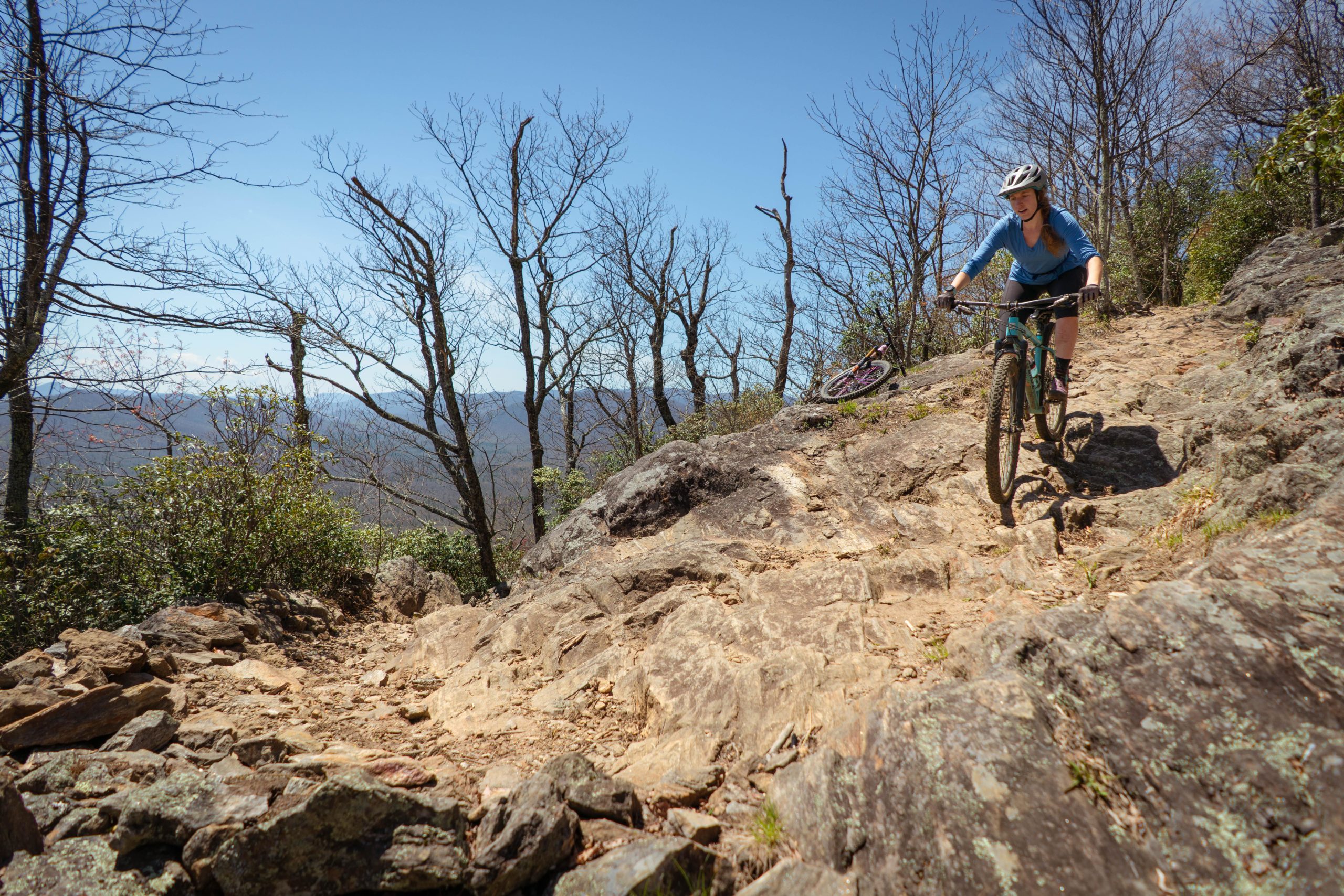 A mountain biker navigating a rocky trail surrounded by sparse trees and distant mountains under a clear blue sky. Pilot Rock mountain bike trail.