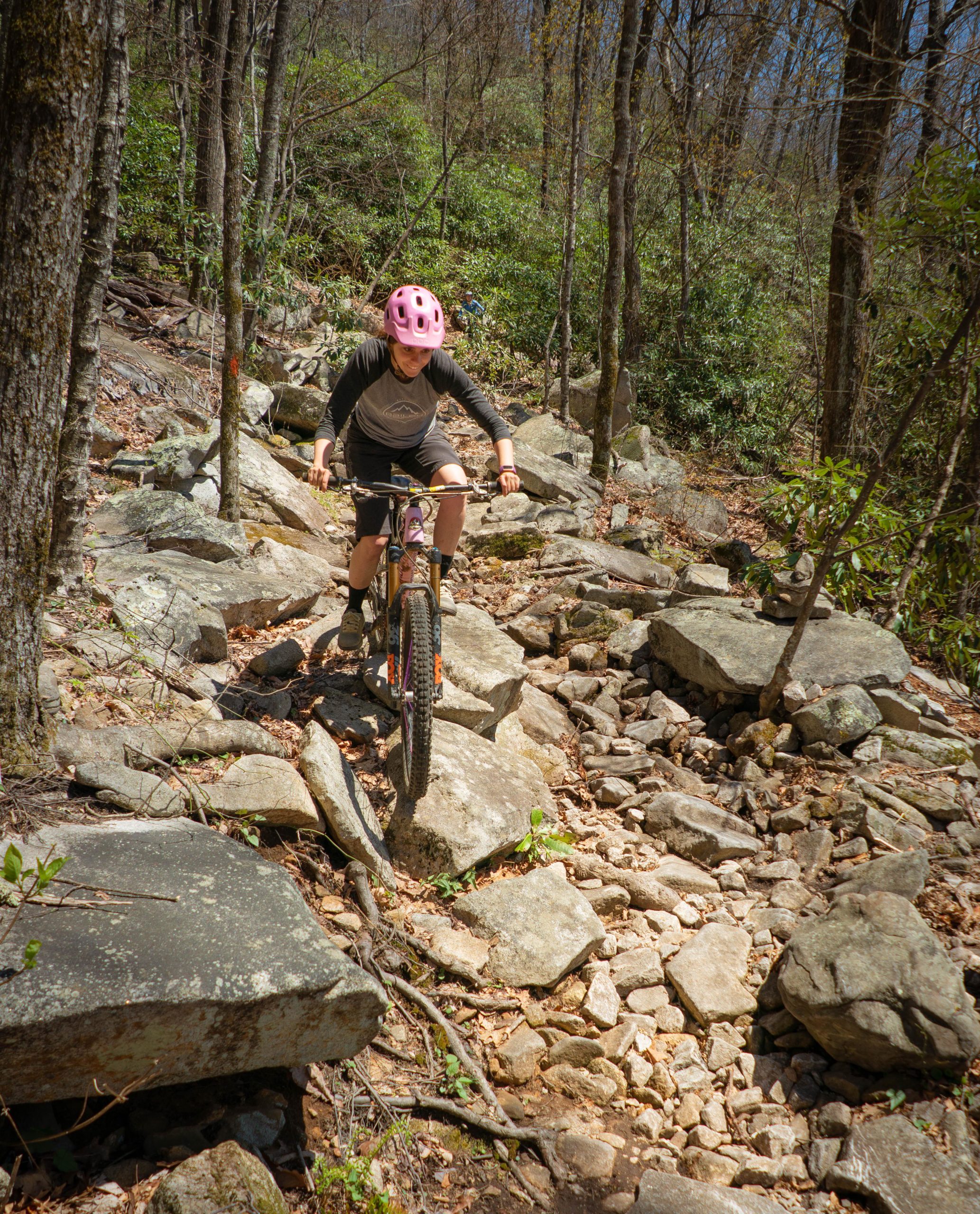 A mountain biker navigating a rocky trail in a wooded area, wearing a pink helmet and a long-sleeve shirt, surrounded by trees and greenery. The terrain consists of large rocks and loose stones, indicating a challenging path for biking. Pilot Rock mountain bike trail.