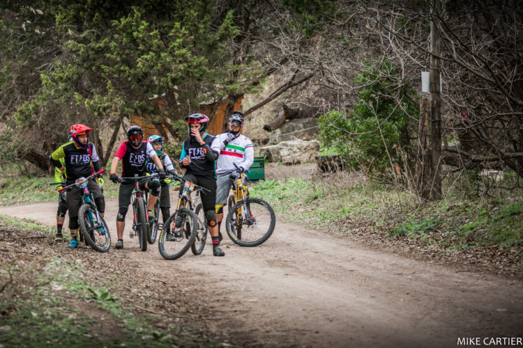 A group of four mountain bikers pausing on a dirt trail surrounded by trees. They are wearing colorful biking gear and helmets, engaging in conversation while resting on their bikes. The scene captures a casual and adventurous moment in nature, with a mix of greenery and earthy tones in the background.