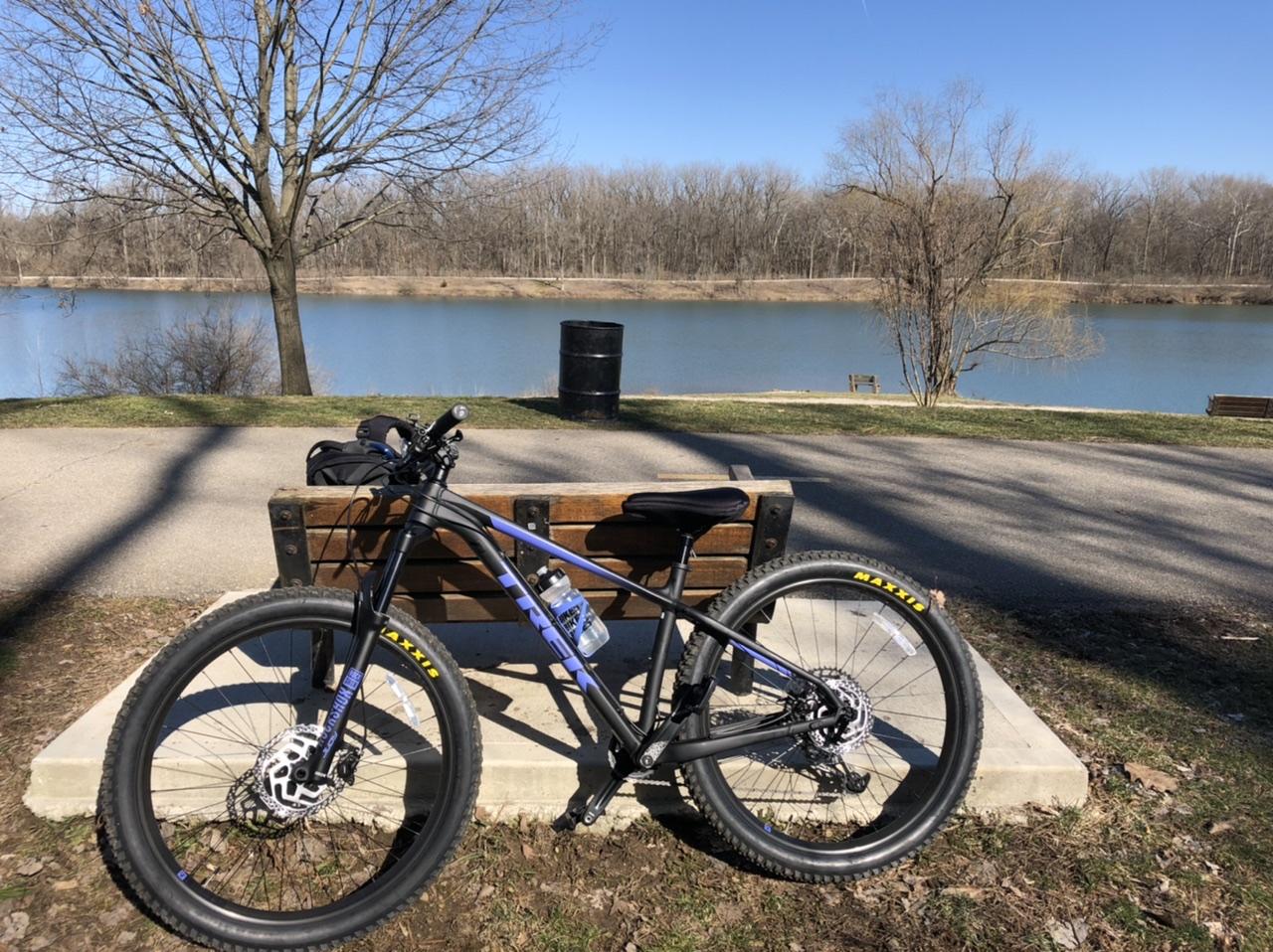 Trek Roscoe 8: A black and blue mountain bike resting on a concrete pad beside a wooden bench, with a serene lake in the background. The scene is set on a sunny day, showcasing clear blue skies and bare trees along the shoreline. A trash can is visible near the lake, and additional benches can be seen in the distance.