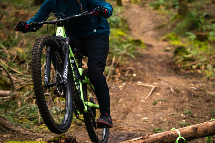 A mountain biker performing a wheelie on a dirt trail in a forested area, surrounded by greenery and fallen logs. The rider is wearing a teal long-sleeve shirt and black pants, with gloves and a focused expression.