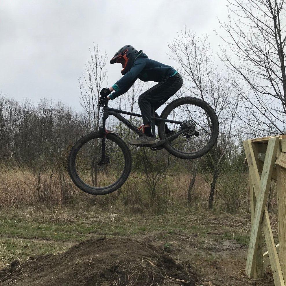A person wearing a helmet and protective gear is mid-air while jumping a mountain bike off a wooden ramp in a natural setting. The background features sparse trees and dry grass under a cloudy sky.
