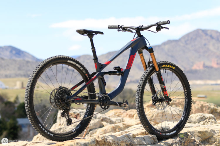 A modern mountain bike is prominently displayed on a rocky surface, with a background of mountains and blue skies. The bike features a sleek design with a dark frame accented by red highlights, and is equipped with thick, knobby tires suitable for off-road terrain. The scene captures a sense of adventure and outdoor activity.