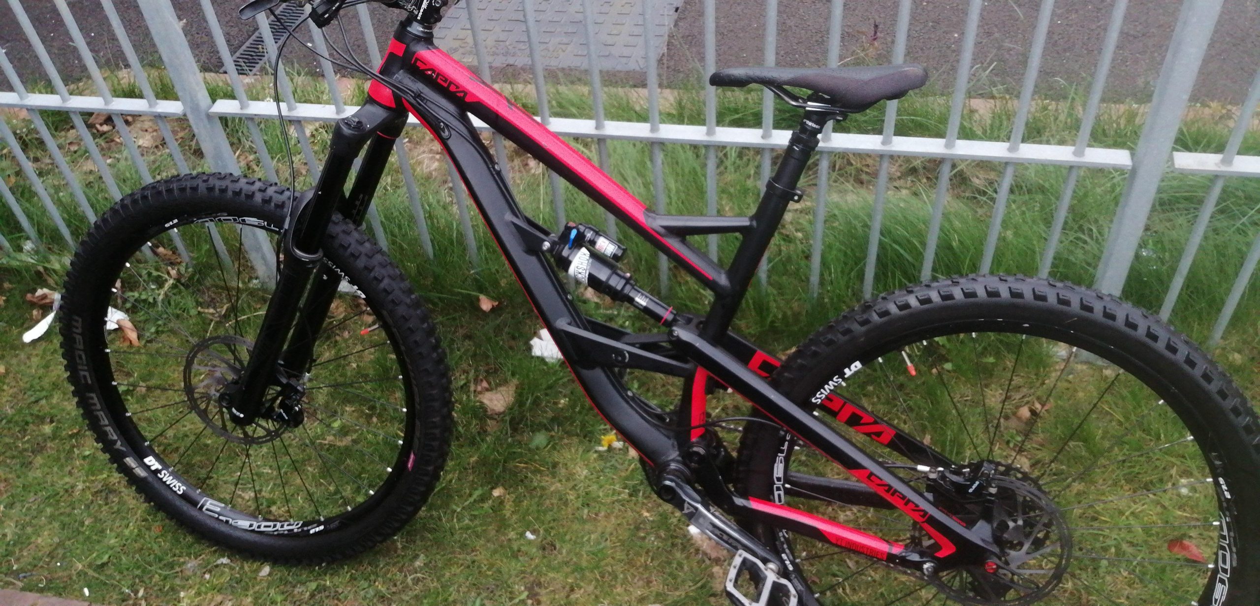 YT Capra: A close-up image of a black and red mountain bike parked next to a fence. The bike features chunky tires, a sturdy frame, and visible suspension components. It is positioned on grass with bits of fallen leaves around.