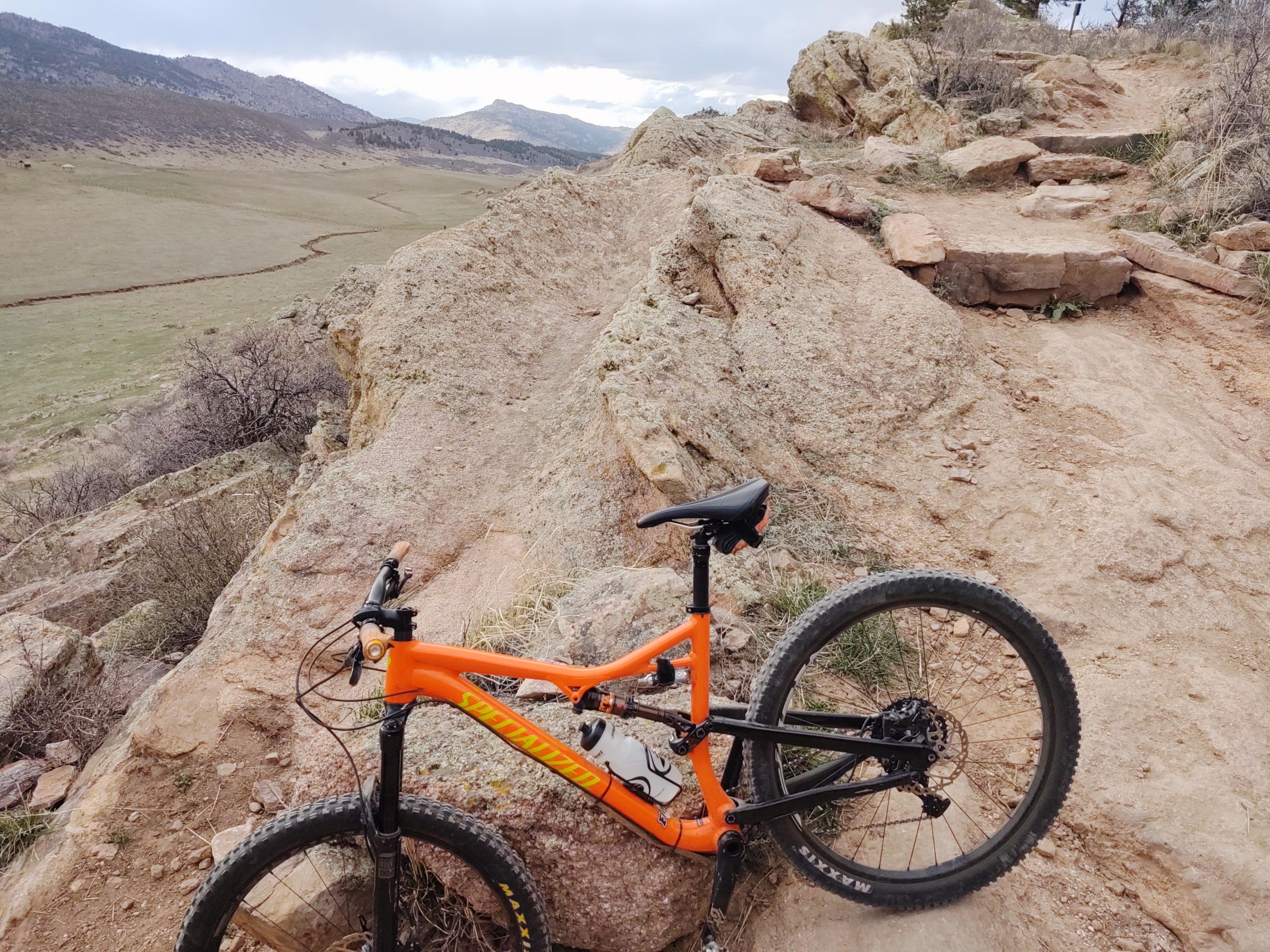 A mountain bike with an orange frame is leaning against a rocky trail, overlooking a grassy valley and distant mountains under a cloudy sky. The terrain features rugged rocks and a narrow dirt path winding through the landscape. Coyote Ridge mountain bike trail.
