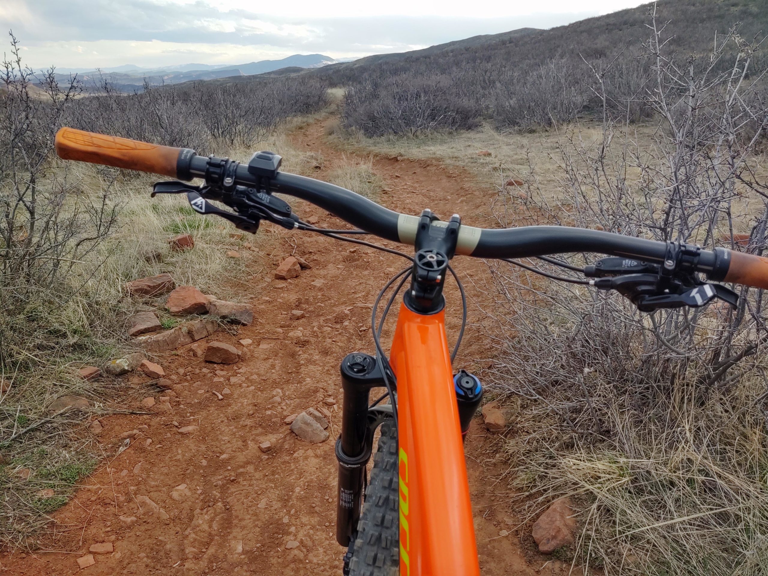 Alt text: A close-up view of mountain bike handlebars and controls, with an orange frame in the foreground. The bike is on a dirt trail surrounded by sparse vegetation and rocky terrain, with distant hills visible in the background under a cloudy sky. Rimrock mountain bike trail.