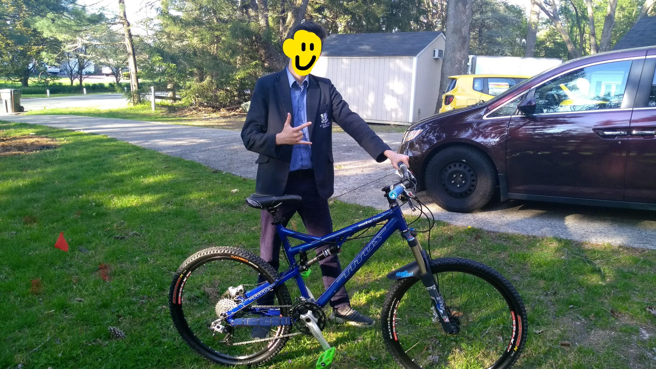 Titus Moto Lite: A person in a jacket stands next to a blue mountain bike, making a hand gesture. The background features a grassy area with trees and parked cars.