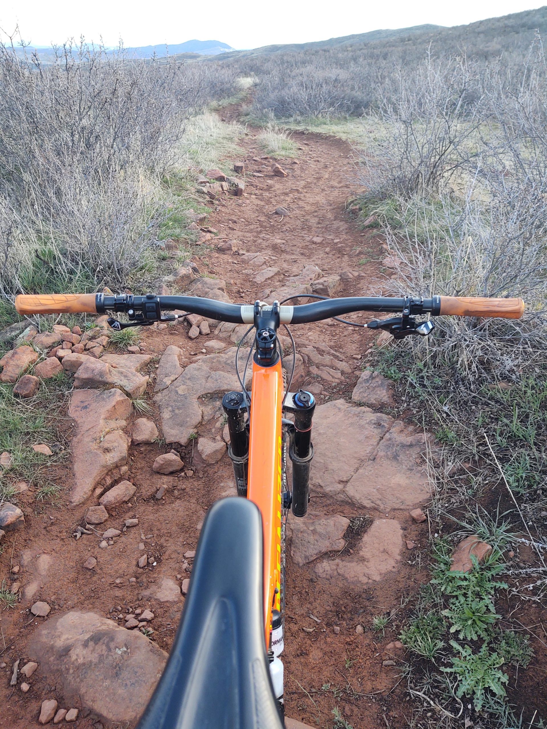 View from the handlebars of a mountain bike positioned on a rocky, dirt trail surrounded by low shrubs and grass, with a distant mountain range visible in the background. Rimrock mountain bike trail.