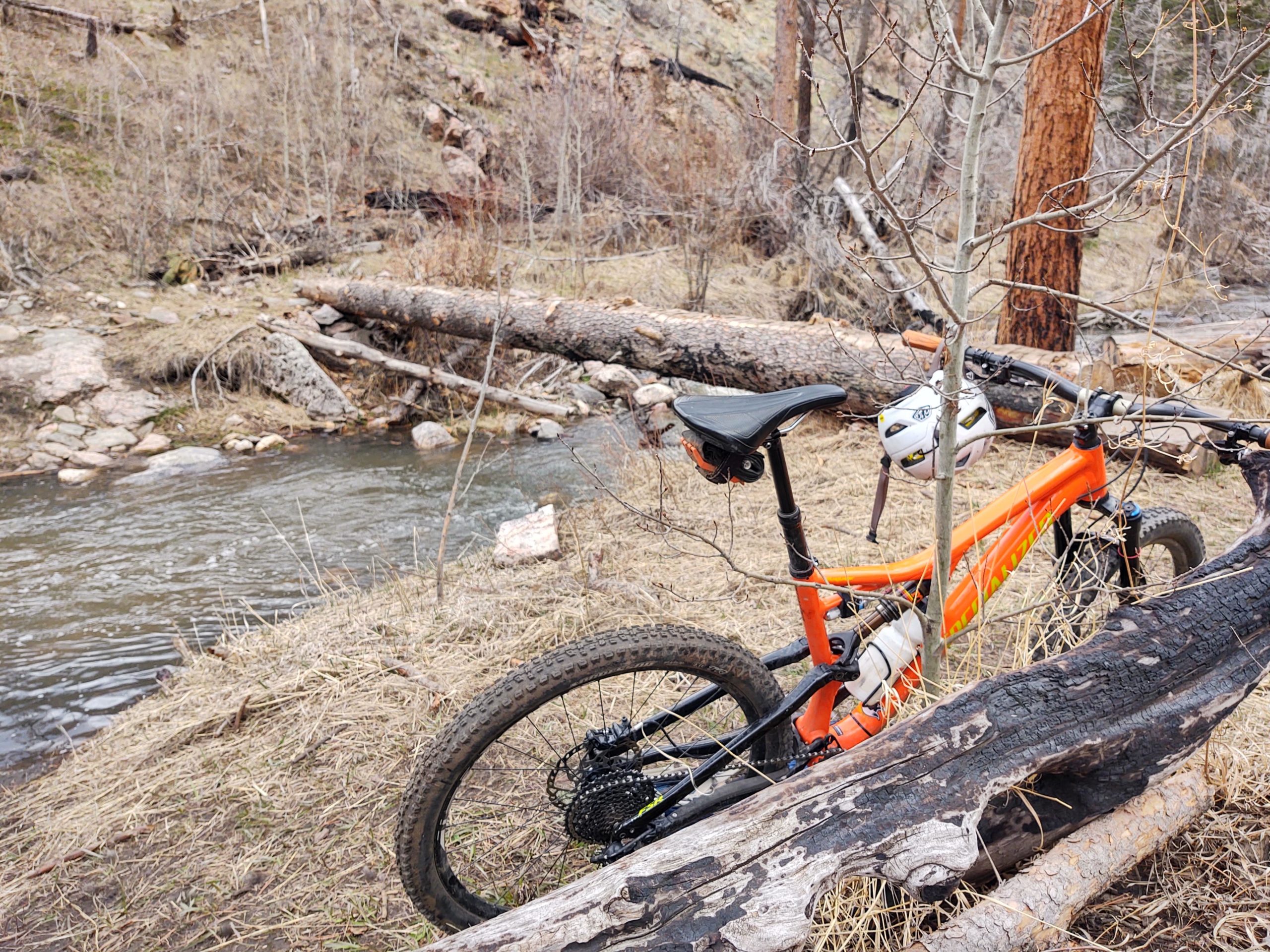 A bright orange mountain bike rests beside a gently flowing stream, surrounded by fallen logs and dried grass. Bare trees dot the landscape, suggesting an early spring environment. The bike is positioned at an angle, with its front wheel partially obscured by a log, and a helmet is mounted on the handlebars. Young Gulch mountain bike trail.