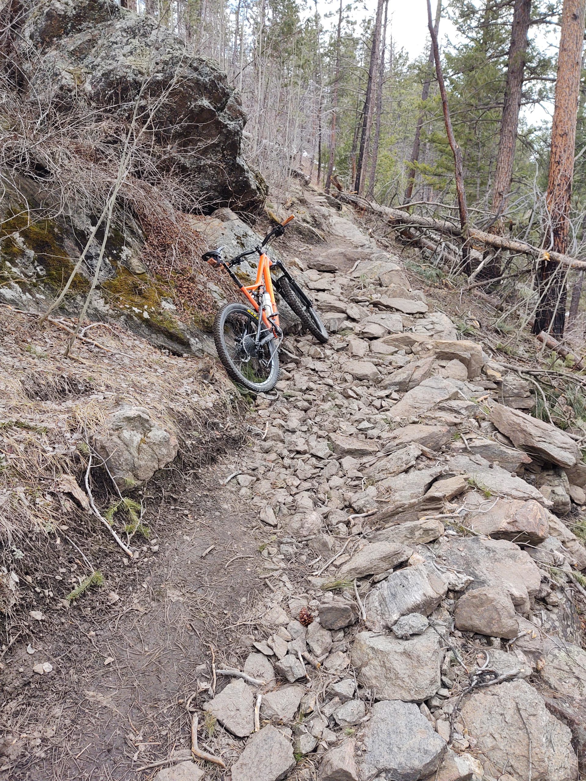 A mountain bike resting against a large rock on a rocky trail surrounded by trees. The path is uneven with scattered stones and roots, indicating a rugged terrain suitable for off-road cycling. Young Gulch mountain bike trail.