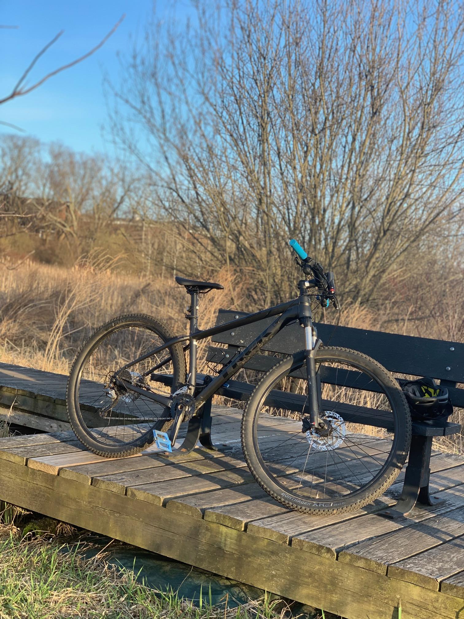 Trek Xcaliber 7: A black mountain bike parked on a wooden bridge with a grassy area in the foreground and bare trees in the background under a clear blue sky.