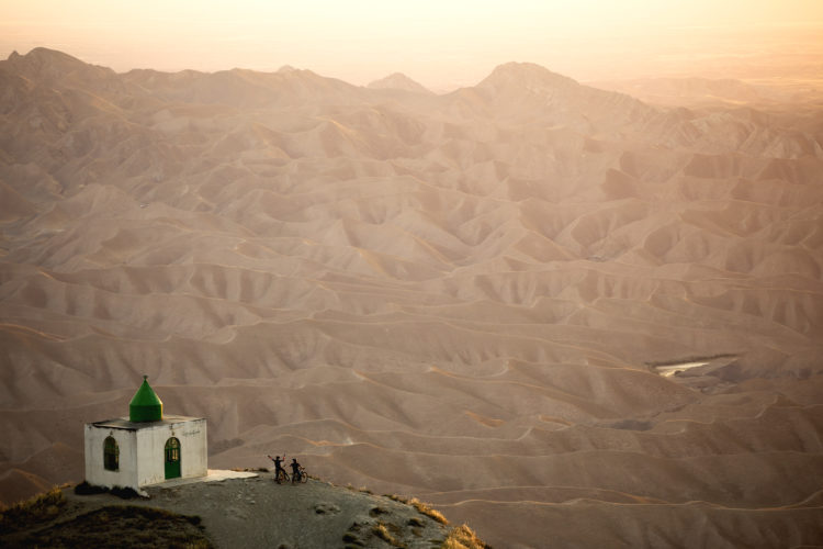 A small white structure with a green dome stands on a hillside, surrounded by rugged brown mountains and valleys in the background. Two individuals with bicycles are seen near the building, enjoying the view during sunset. The landscape features soft, wavy patterns of earth, illuminated by warm golden light.