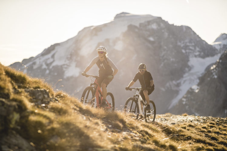 A vibrant scene of two mountain bikers navigating a rocky trail surrounded by majestic mountains. The sun is setting in the background, casting a warm glow over the landscape, highlighting the rugged terrain and grassy patches. One biker is on a red mountain bike, while the other rides a black bike, both wearing helmets and athletic gear.