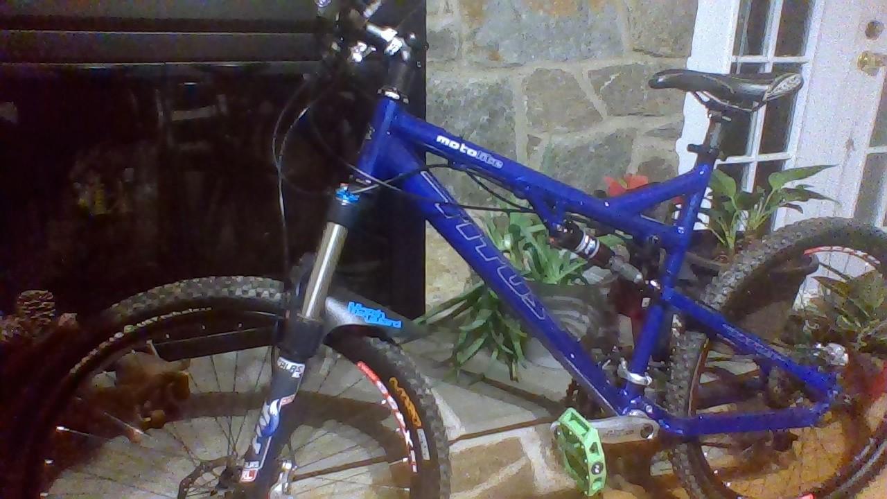 Titus Moto Lite: A blue mountain bike positioned indoors, leaning against a stone wall. The bike features large tires, a sturdy suspension fork, and colorful pedals. In the background, there are decorative plants and a glass-door entryway.