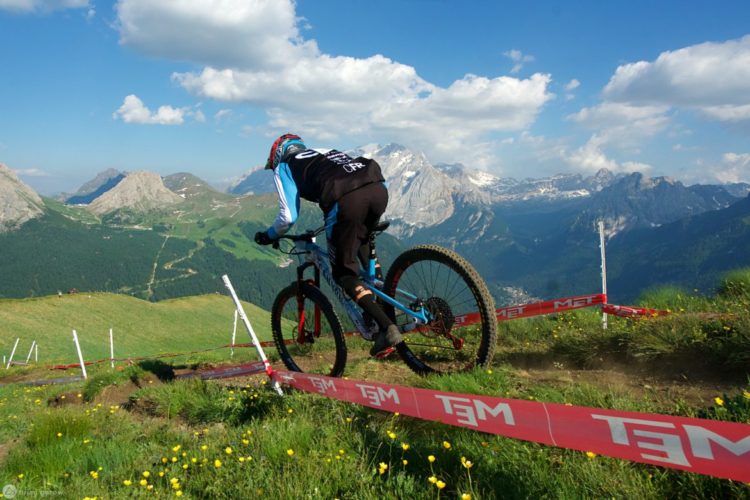 A mountain biker navigating a winding trail on a lush green hillside, with snow-capped mountains in the background. The rider is wearing protective gear and leaning forward on a blue and black mountain bike, with red barrier tape marking the course. The sky is partly cloudy, adding to the scenic mountain landscape.