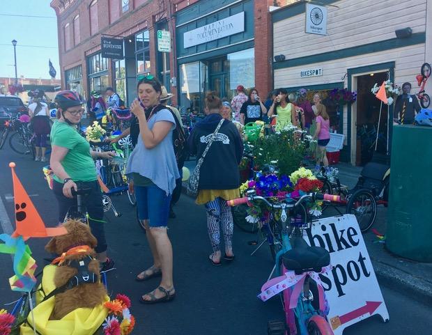 A lively street scene featuring a group of people gathered around a colorful bike decoration event. Several bicycles are adorned with flowers and festive decorations, while participants, including women wearing casual attire and helmets, interact and enjoy the atmosphere. A sign reading "bike spot" is visible, and the backdrop includes shops and storefronts.