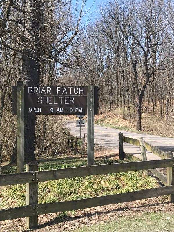 Sign for Briar Patch Shelter, indicating it is open from 9 AM to 8 PM, with a wooded path and road visible in the background. Bonneyville Mill mountain bike trail.