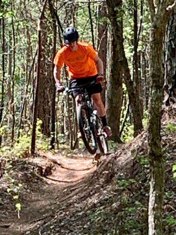 A mountain biker in an orange shirt jumps off a dirt path in a forested area, surrounded by trees and greenery. The rider is wearing a helmet and appears to be focused as they navigate the trail. Fort Yargo State Park mountain bike trail.