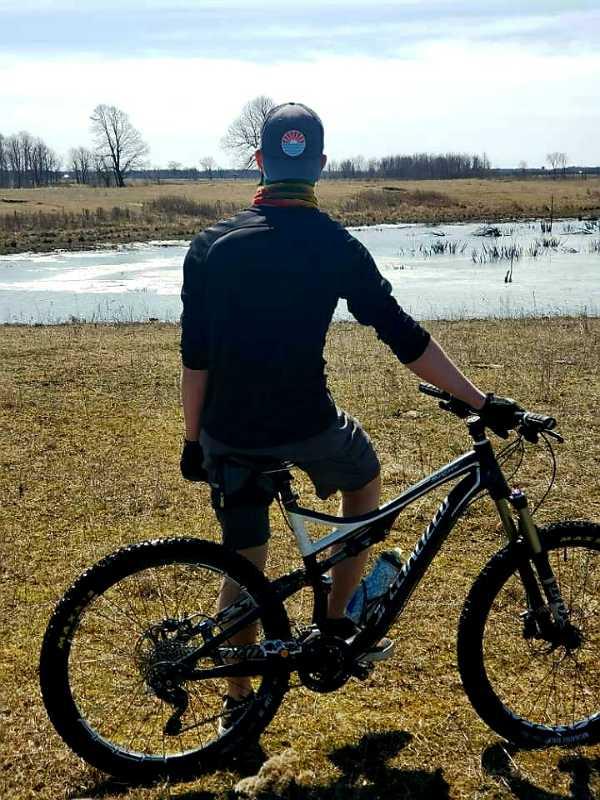 Person standing beside a mountain bike, looking out over a grassy field and water. The scene is set on a sunny day, with a blue sky and a few trees in the background. The cyclist is wearing a cap and a colorful neck gaiter, dressed in a black long-sleeve shirt and shorts. Baie des Atocas Trail mountain bike trail.