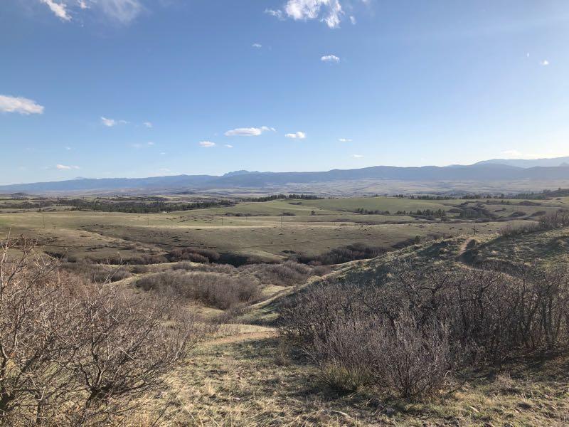 A panoramic view of rolling hills and valleys under a clear blue sky, with scattered clouds. The foreground features dry shrubs and grasses, while the distance reveals lush fields and mountains on the horizon. Highlands Point mountain bike trail.