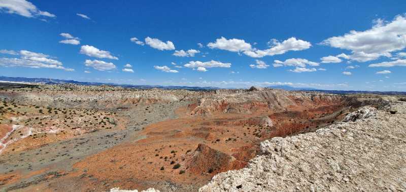 A panoramic view of a rugged, colorful landscape featuring layers of red and gray earth, with sparse vegetation and a dramatic blue sky dotted with white clouds. The terrain shows various geological formations and valleys, capturing the essence of a remote, arid environment. White Ridge Bike Trails mountain bike trail.