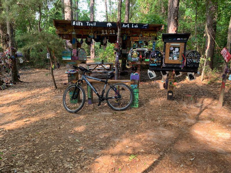 A mountain bike is parked in front of a colorful art installation in a wooded area. The structure features various painted signs and decorations, including a roof sign that reads "Bill's Trail Art Lab." Surrounding the art lab are tall trees and a dirt path, providing a tranquil outdoor setting. Bill's Trail mountain bike trail.