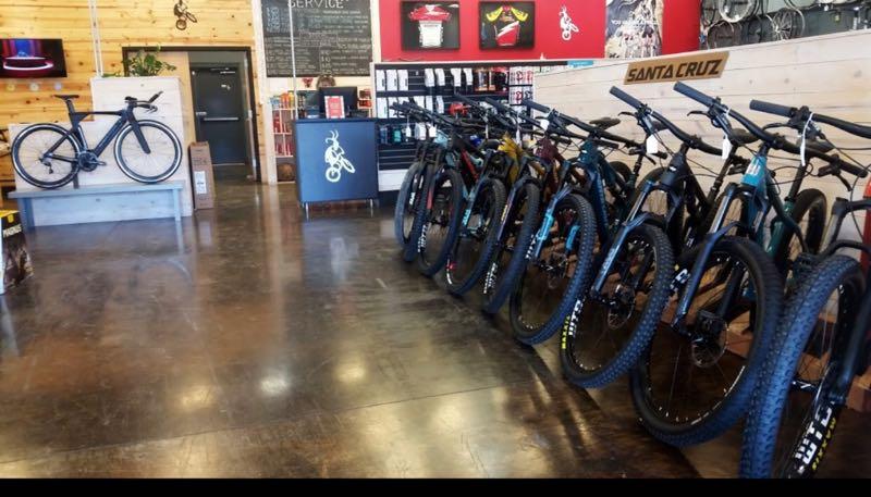 Bicycle shop interior featuring a row of mountain bikes lined up against a wall, with a large black bike displayed on a pedestal in the foreground. The shop has wooden walls, a service area in the background, and various biking gear displayed on shelves.
