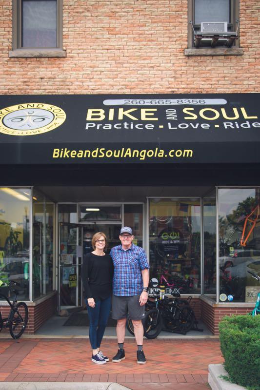 A man and a woman stand together in front of the "Bike and Soul" shop, a bicycle store with a black awning displaying their logo and the tagline "Practice, Love, Ride." The storefront features large glass windows showcasing various bicycles and accessories. The pair are smiling, dressed casually, with the shop's contact information and website visible above the entrance.