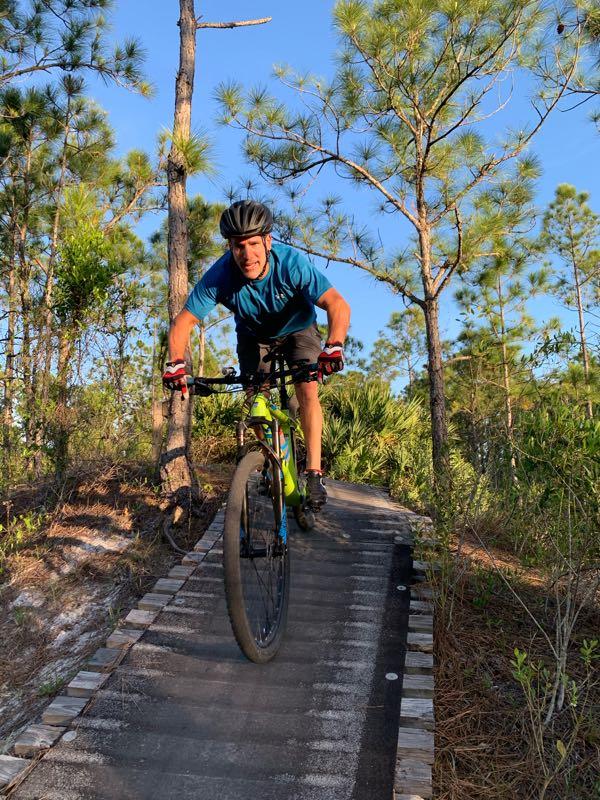 A cyclist in a blue shirt and helmet rides a mountain bike along a wooded trail, leaning forward as they navigate a narrow wooden path surrounded by green trees and foliage. The sun shines in a clear blue sky, creating a vibrant outdoor scene. Halpatiokee mountain bike trail.