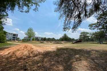 A sunny park landscape featuring a dirt bike area with ramps and mounds, surrounded by grass and trees. In the background, there are several residential buildings and a clear blue sky with a few clouds. Orlando Mountain Bike Park mountain bike trail.