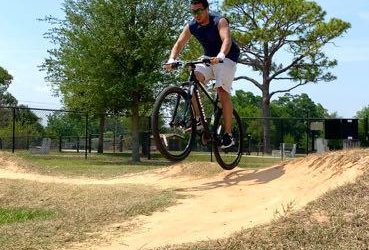 A person riding a mountain bike, airborne while executing a jump on a dirt track in a park. Surrounding the track are green trees and grass under a clear blue sky. Orlando Mountain Bike Park mountain bike trail.