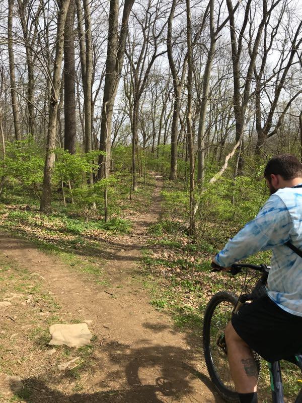 A person in a tie-dye shirt is standing next to a mountain bike on a dirt path winding through a lush green forest. The path is flanked by tall, bare trees and small green shrubs, indicating early spring foliage. Jordan Creek Parkway mountain bike trail.