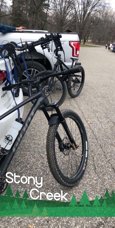 A close-up view of two mountain bikes parked near a pickup truck on a gravel road at Stony Creek. The bikes are positioned at an angle, showcasing their tires and handlebars, with trees in the background indicating a natural setting. The image features text at the bottom labeled "Stony Creek" with a green forest-themed border. Stony Creek Metro Park mountain bike trail.