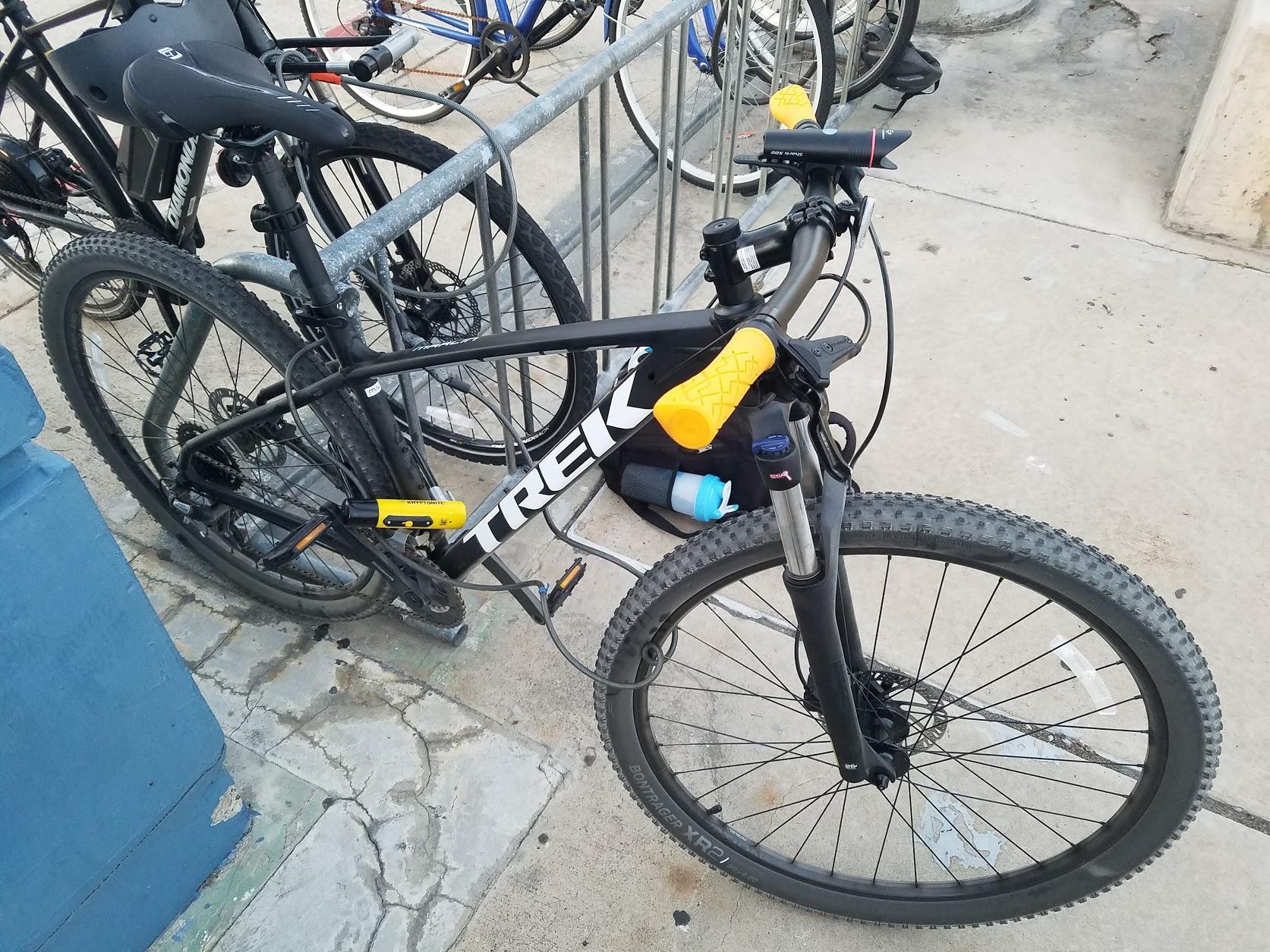 Trek Marlin 5: A black Trek mountain bike is parked at a bike rack. It features yellow handle grips and a U-lock secured through the frame. In the background, more bicycles are visible, and the ground has a cracked concrete surface.