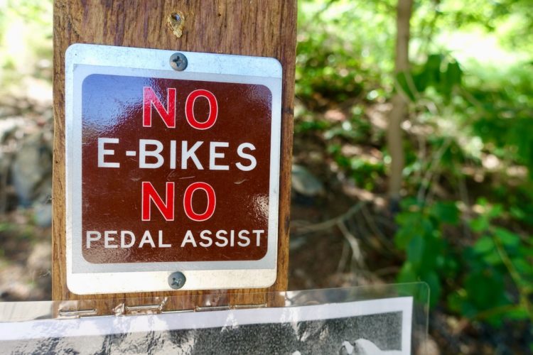 A wooden post with a sign reading "NO E-BIKES NO PEDAL ASSIST" in bold red letters. The background features blurred greenery, indicating a natural outdoor setting.