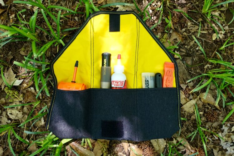 A partially opened tool pouch featuring a bright yellow interior with various tools and supplies organized inside. Items include a black marker, a small container, a bottle of sealant, and a bright orange tool, all resting on a bed of grass and leaves.