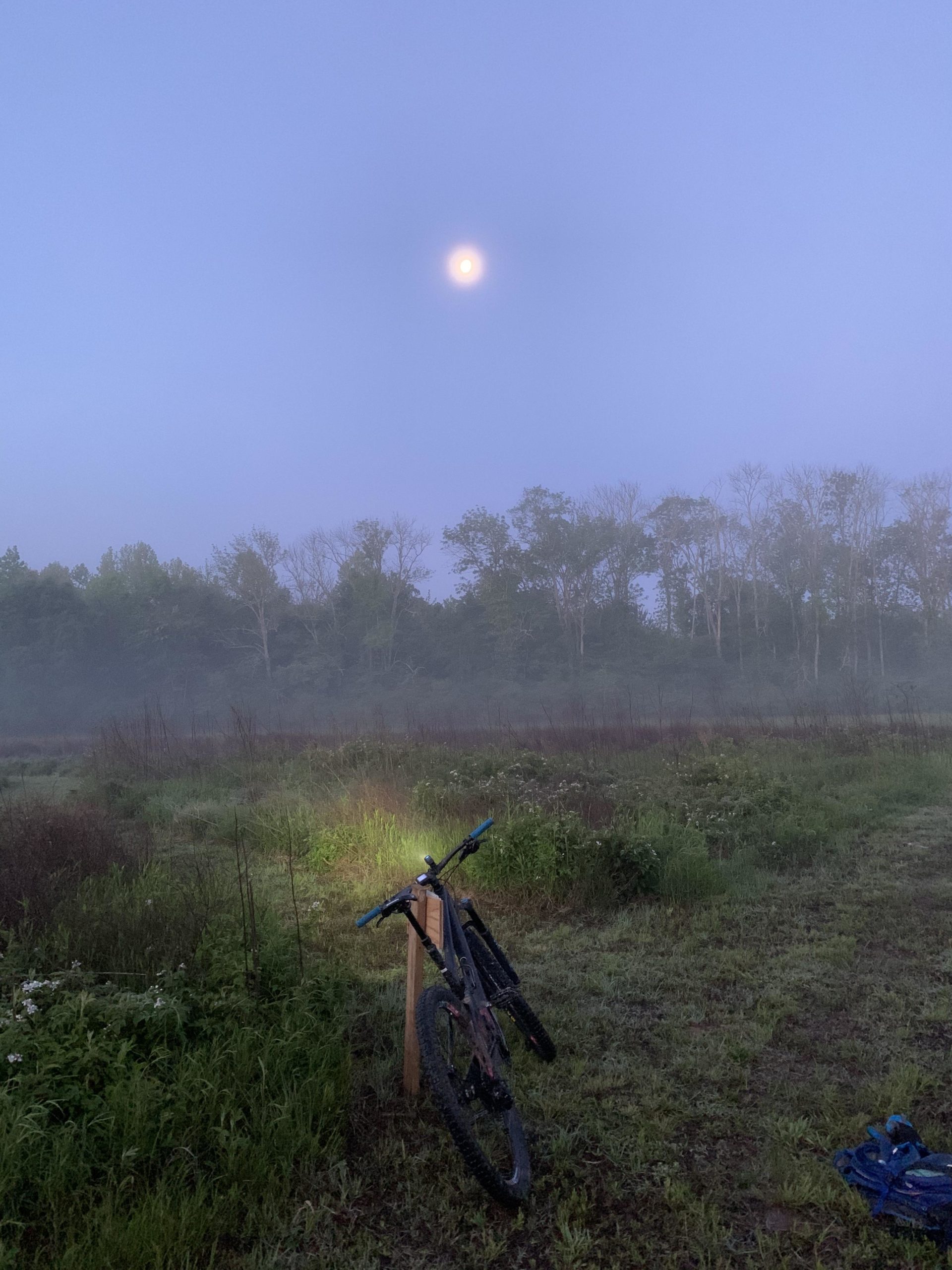 A mountain bike resting against a wooden post in a misty field at dawn, with a bright full moon visible in a cloudy sky above. The landscape is surrounded by green grass and sparse trees, enhancing the serene and tranquil atmosphere of the early morning. Allatoona Creek Park mountain bike trail.
