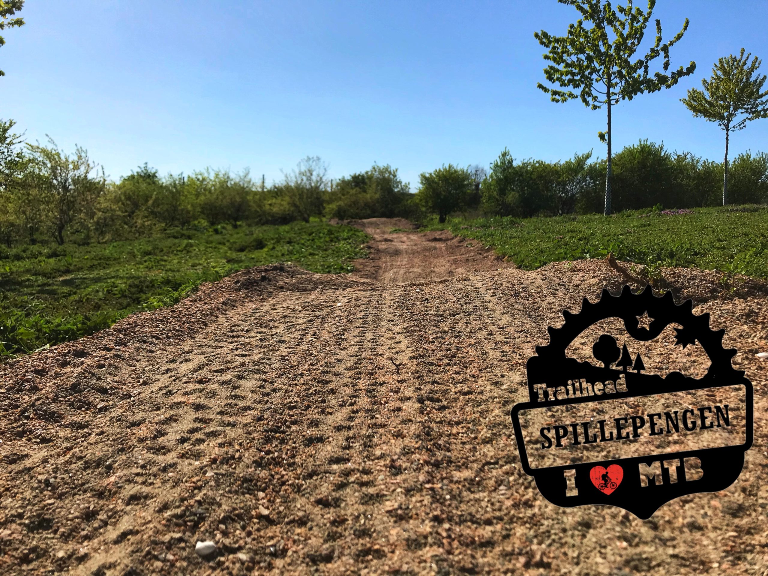 A dirt trail winding through a green landscape with trees and blue sky, featuring a logo in the corner that reads "Trailhead Spillepengen I ❤️ MTB." The trail