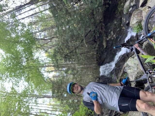 A person wearing a helmet and gloves stands beside a mountain bike near a flowing stream in a lush, green forest. The backdrop features trees with fresh foliage and a rocky area alongside the water. Bear Creek mountain bike trail.