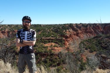 A person wearing a skeleton-printed cycling jersey and a black helmet stands with crossed arms on a rocky outcrop overlooking a canyon with red cliffs and sparse greenery under a clear blue sky. Caprock Canyons Trail System mountain bike trail.