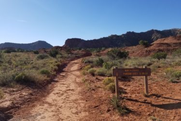 A dirt trail winding through a rocky landscape with sparse vegetation, leading towards a sign that reads "North Prong Parking Area 13 Miles." The background features rugged hills and a clear blue sky. Caprock Canyons Trail System mountain bike trail.