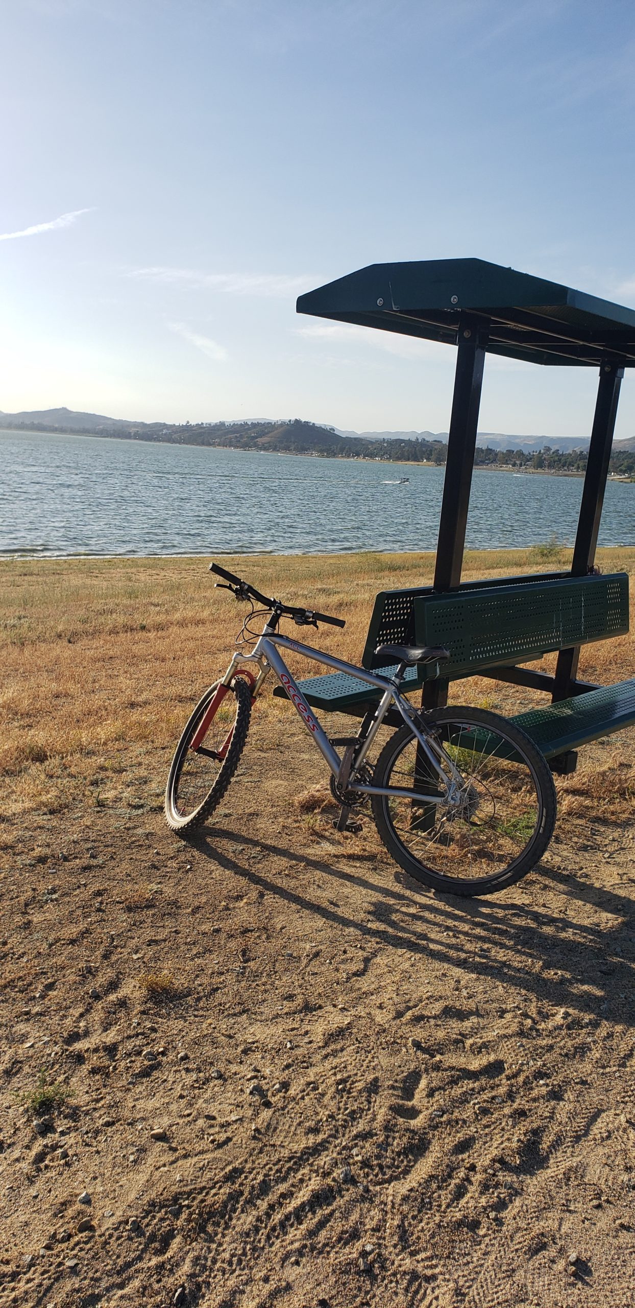 A bicycle leaning against a bench by a lake under a clear blue sky, with hills visible in the distance and dry grass in the foreground. Lake Elsinore mountain bike trail.