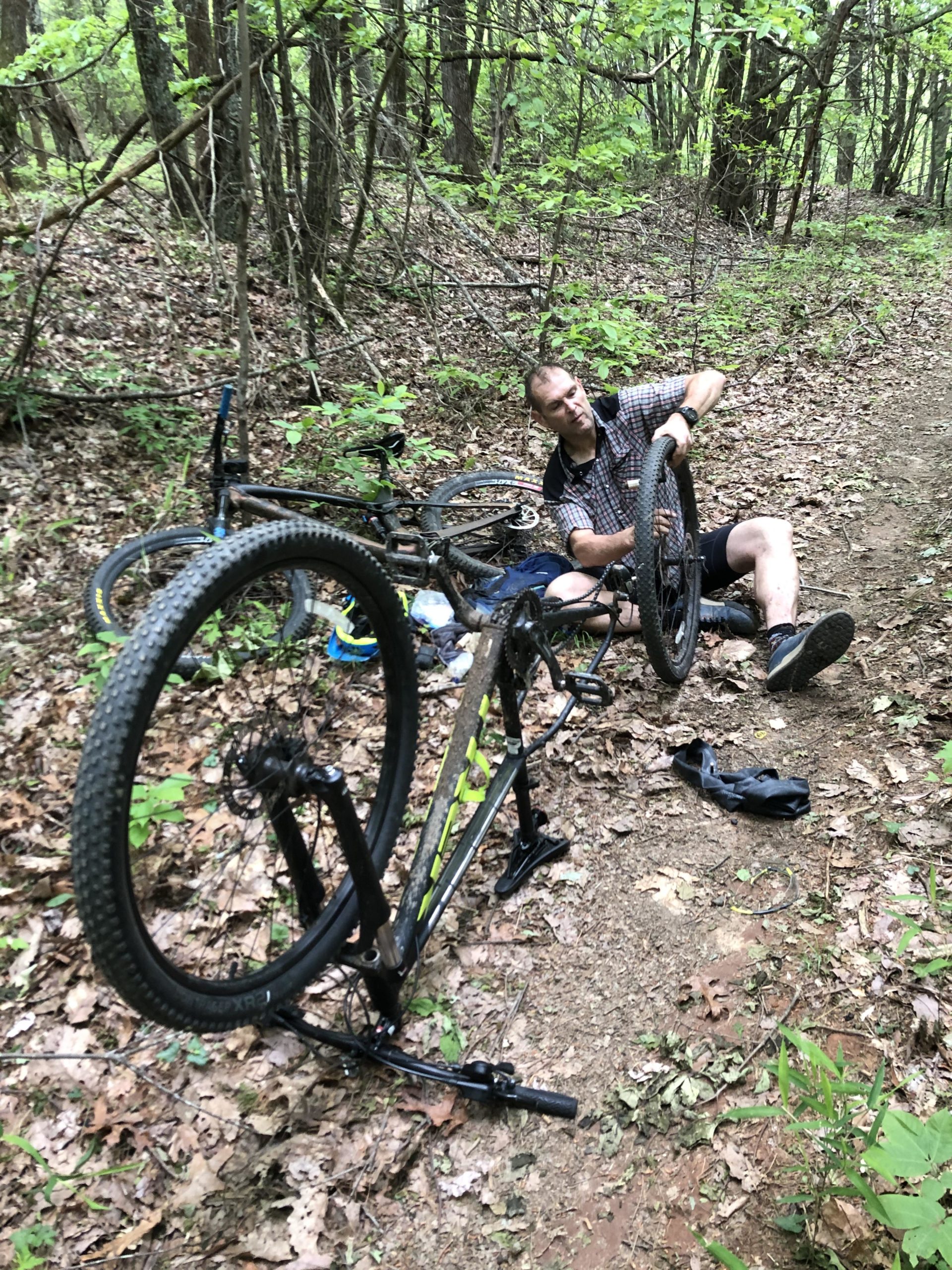 A person sitting on the ground in a forested area, working on a bicycle. The bicycle is propped upside down, with one wheel in the person's hands. Surrounding them are trees and underbrush, creating a natural setting with fallen leaves and greenery. Bull / Jake Mountain mountain bike trail.