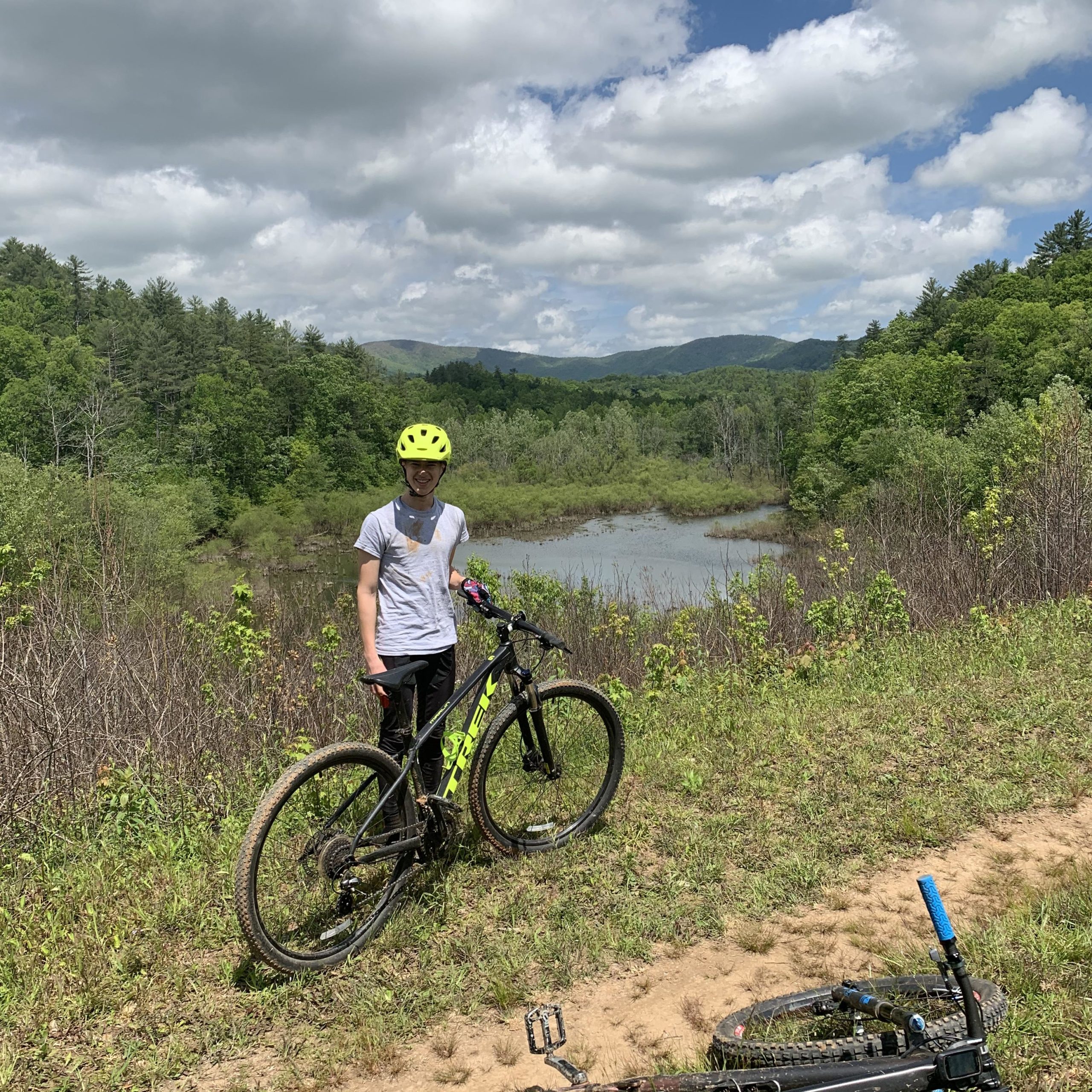 A person wearing a yellow helmet stands next to a mountain bike on a grassy hillside overlooking a calm body of water, surrounded by lush green trees and mountains under a partly cloudy sky. Bull / Jake Mountain mountain bike trail.