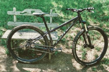 A mountain bike parked next to a wooden bench on a grassy area, with sunlit greenery in the background. The bike features thick, knobby tires and a sturdy frame, suggesting it's ready for outdoor trails. Great Bear mountain bike trail.