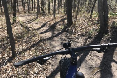 A perspective view from the handlebars of a mountain bike, showing a winding dirt trail through a forest of tall trees. Sunlight filters through the leaves, illuminating the path covered in fallen leaves and patches of grass. Glacial Blue Hills mountain bike trail.