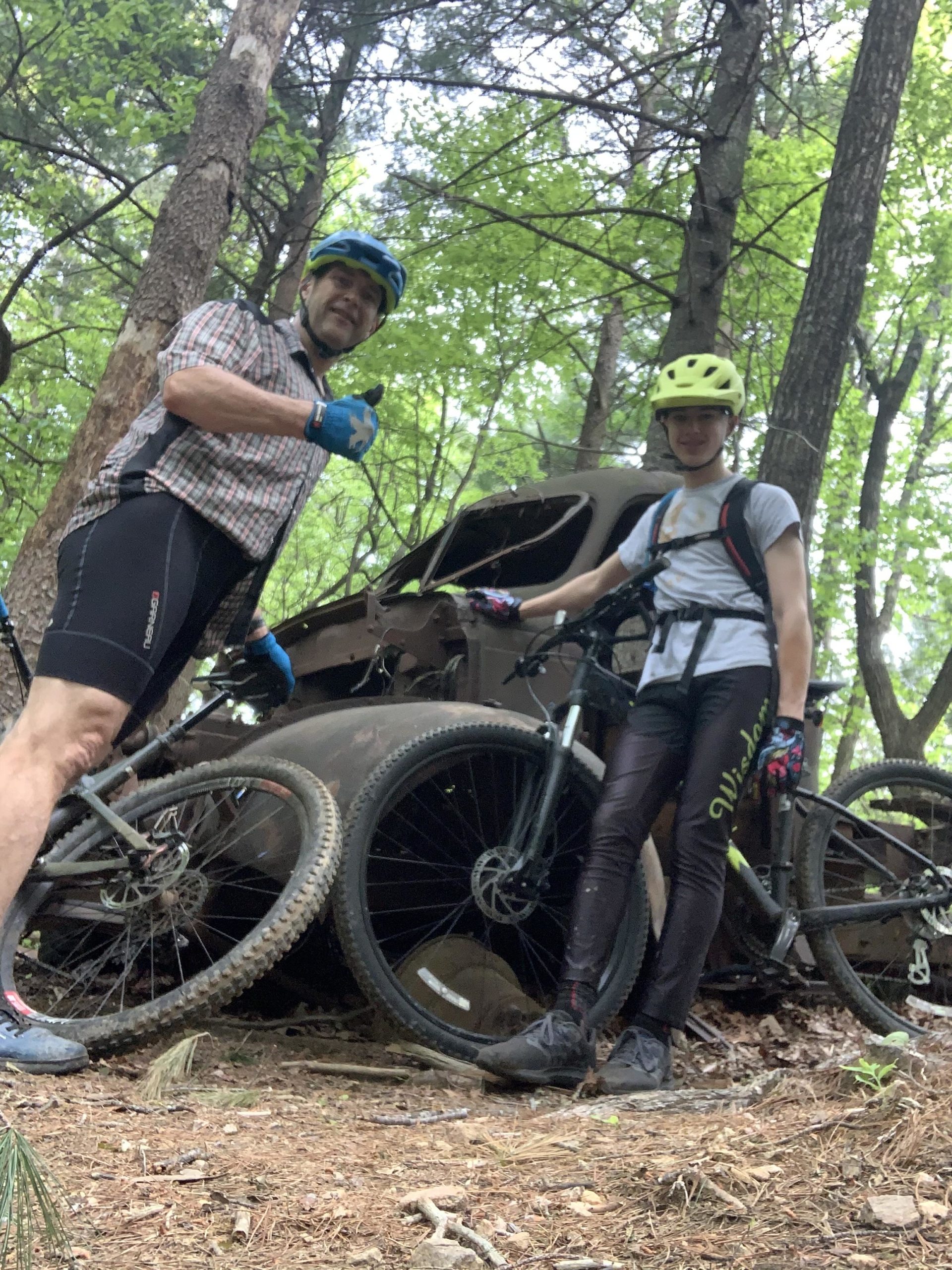 Two individuals in cycling gear pose beside an old, rusted vehicle in a wooded area. The person on the left gives a thumbs-up, while the person on the right stands with their bike, both smiling. Lush green trees and a dirt path are visible in the background. Bull / Jake Mountain mountain bike trail.
